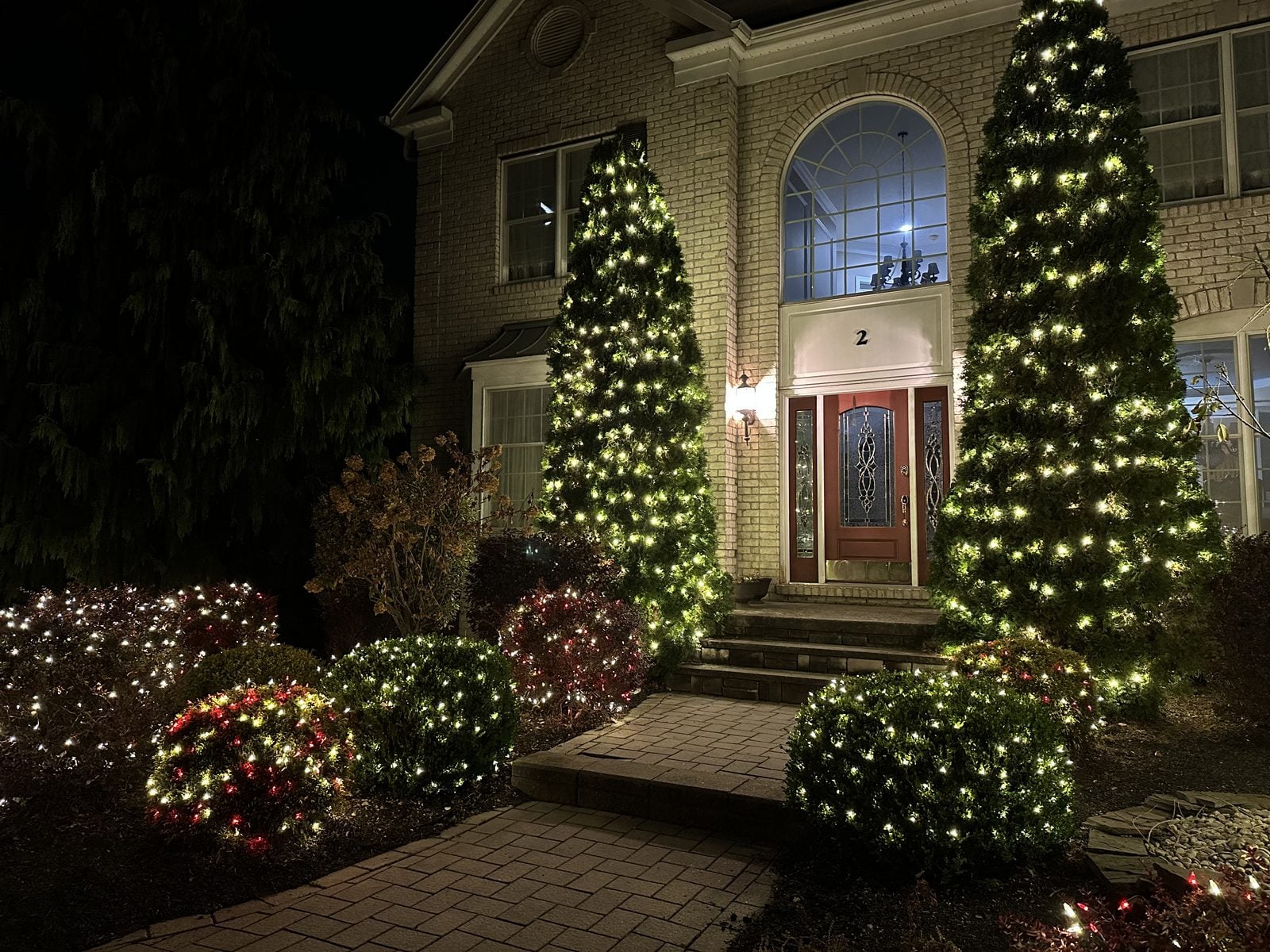 A front entry flanked by two tall lit Christmas trees, garland running over the door