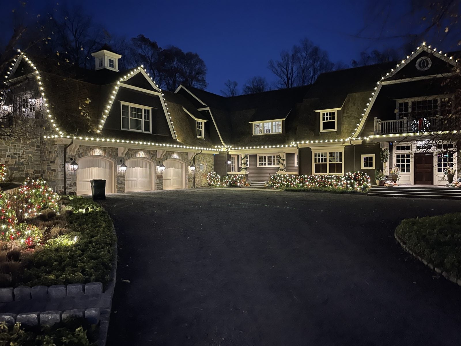 A shingled estate with warm bulbs along the eaves and wreaths on the garage bays