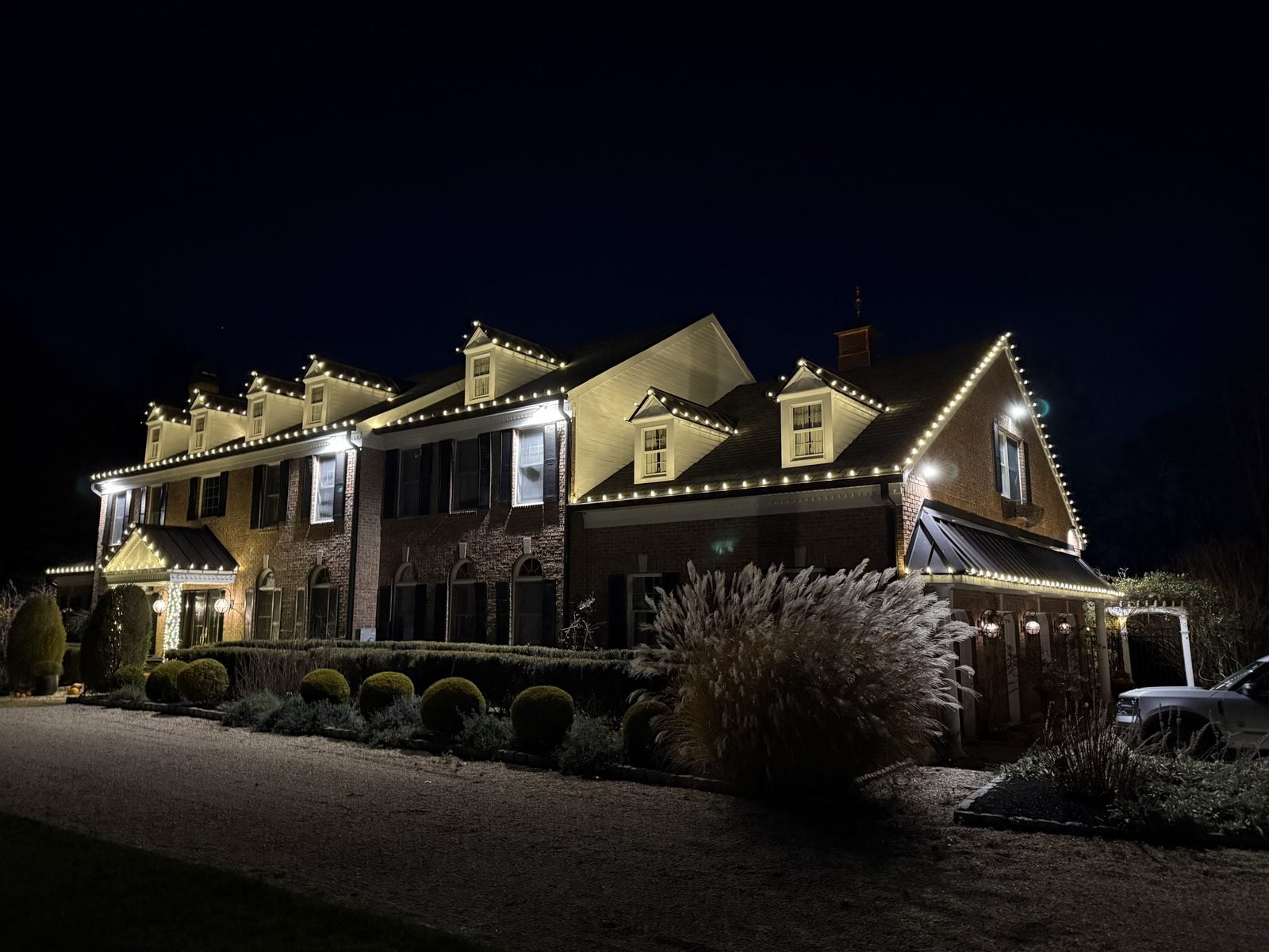 A shingled house with warm bulbs along the eaves and lit planting below