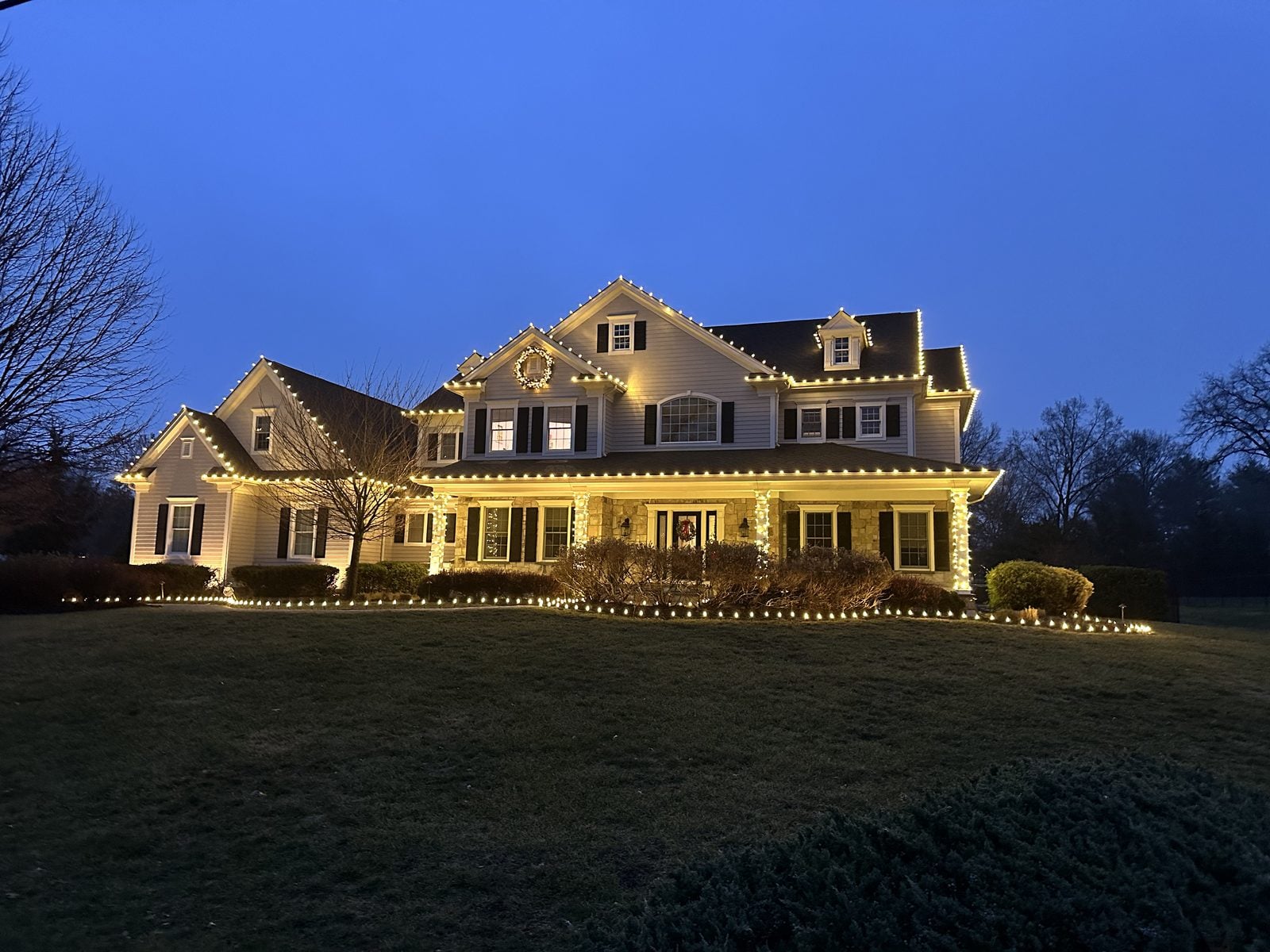 A colonial home at blue hour with warm bulbs along every roofline and garland at the windows