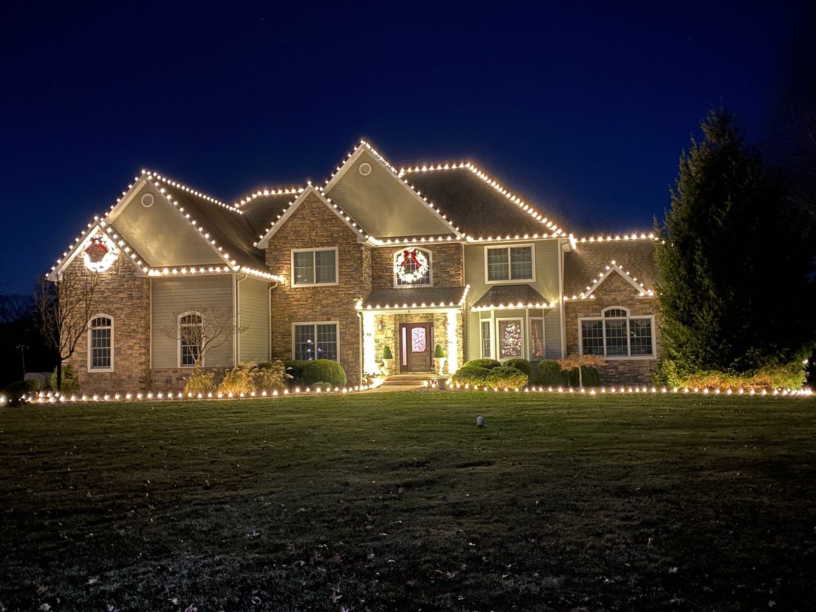A brick colonial outlined in warm bulbs with lit wreaths at every window