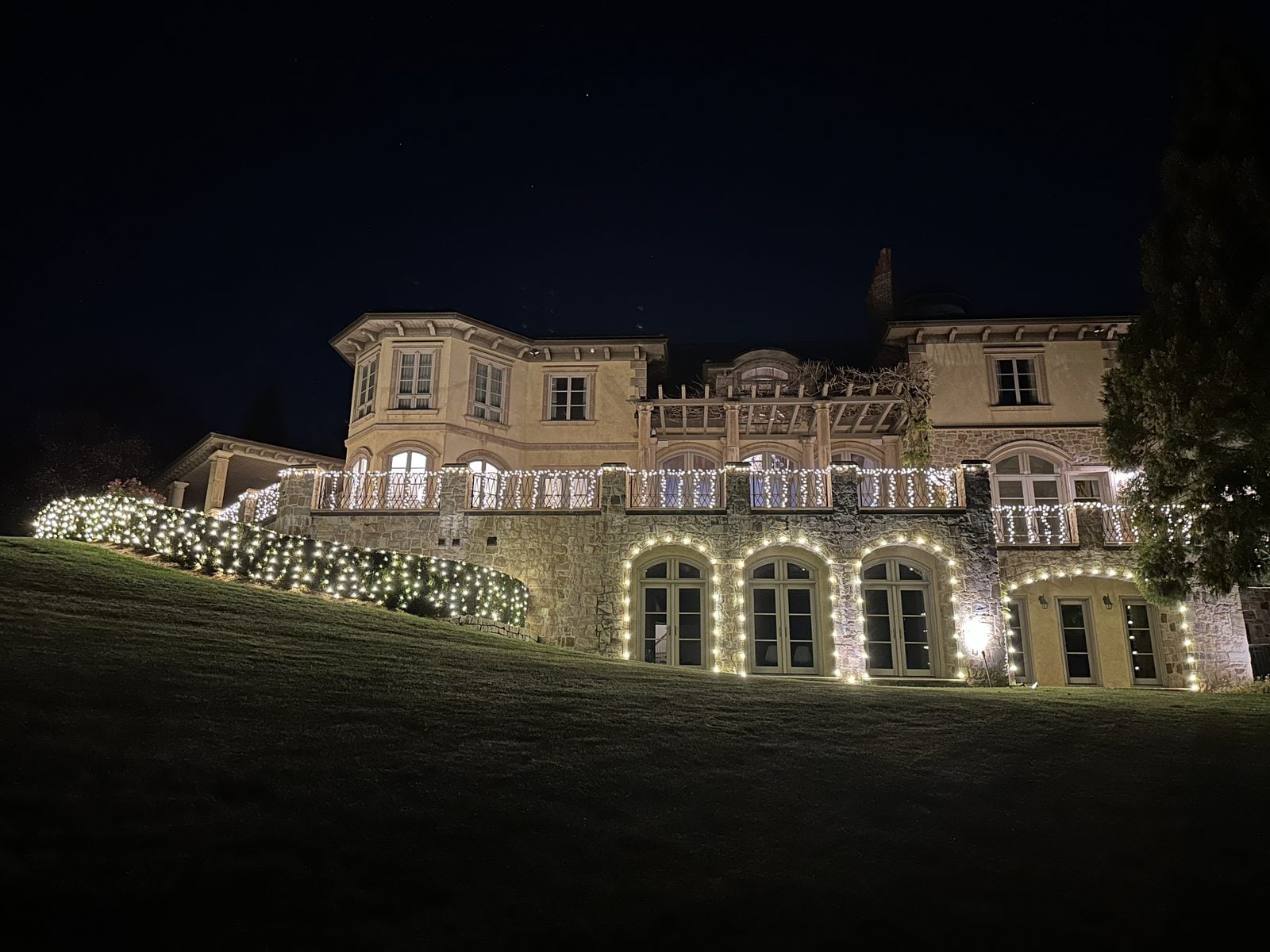 A large stone home with roofline lighting and lit planting along the foundation