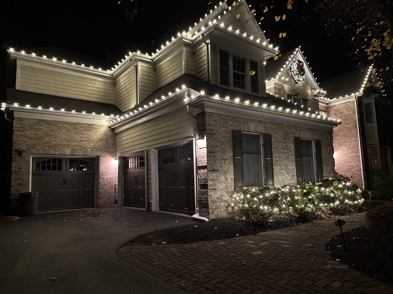 Garage bays outlined in warm bulbs with lit shrubs and a wreath above the doors