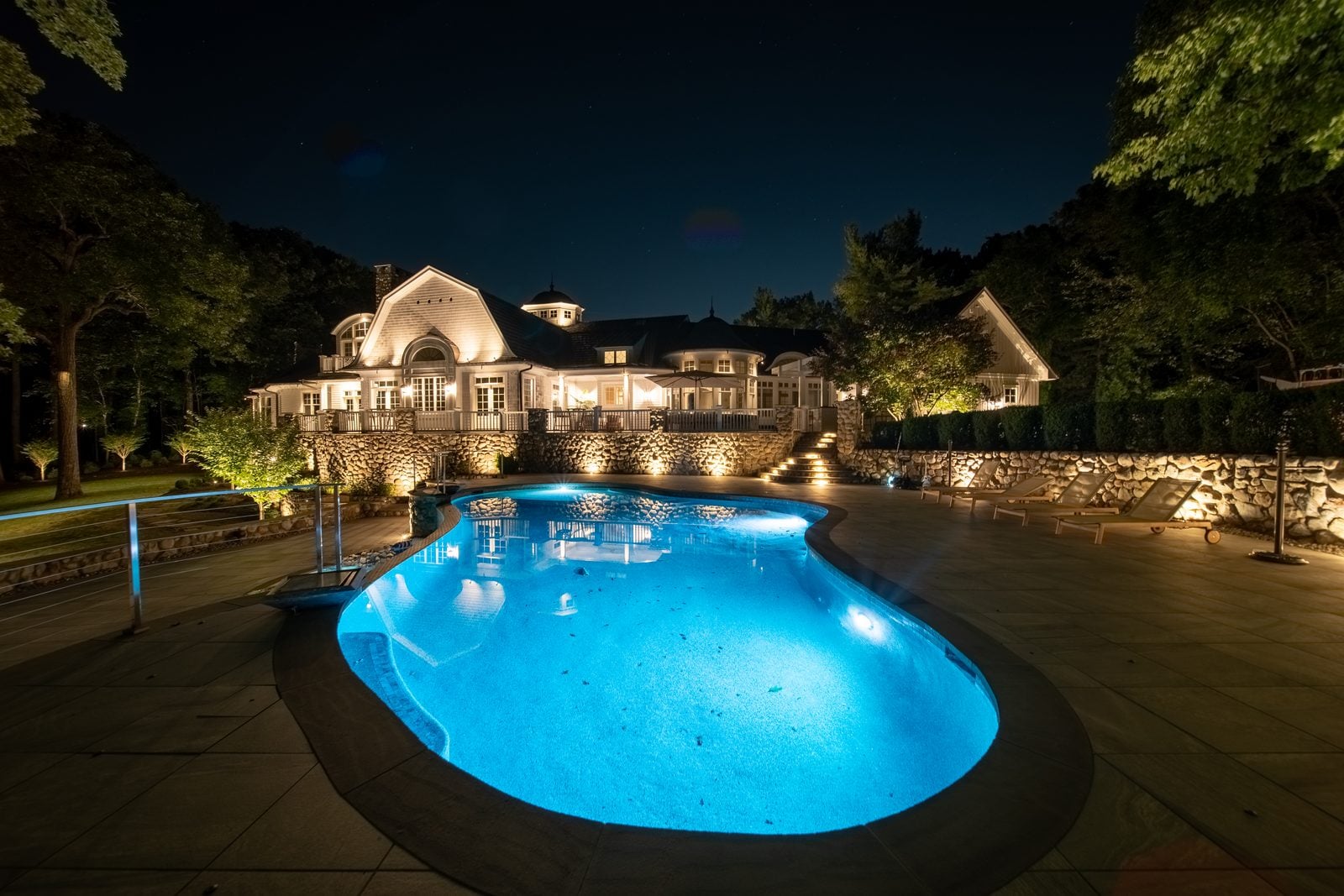 A free-form pool lit from within beside a shingled house, warm light on the stone terrace