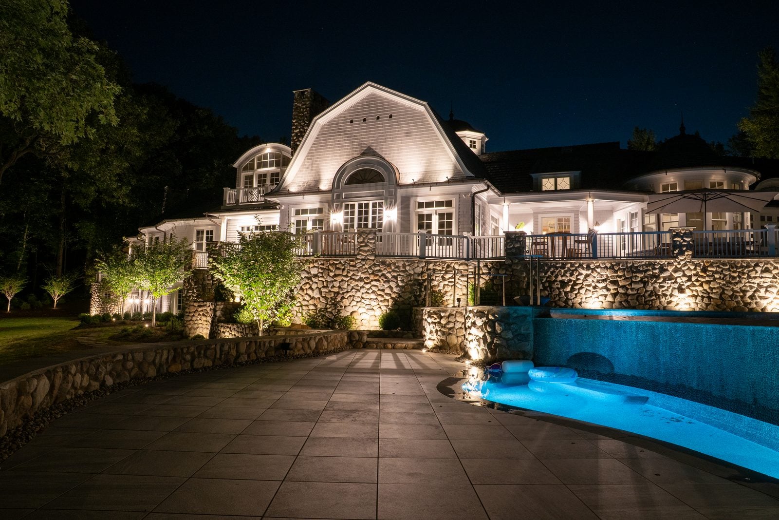 A pool and terrace lit after dark, the far planting uplit above the waterline