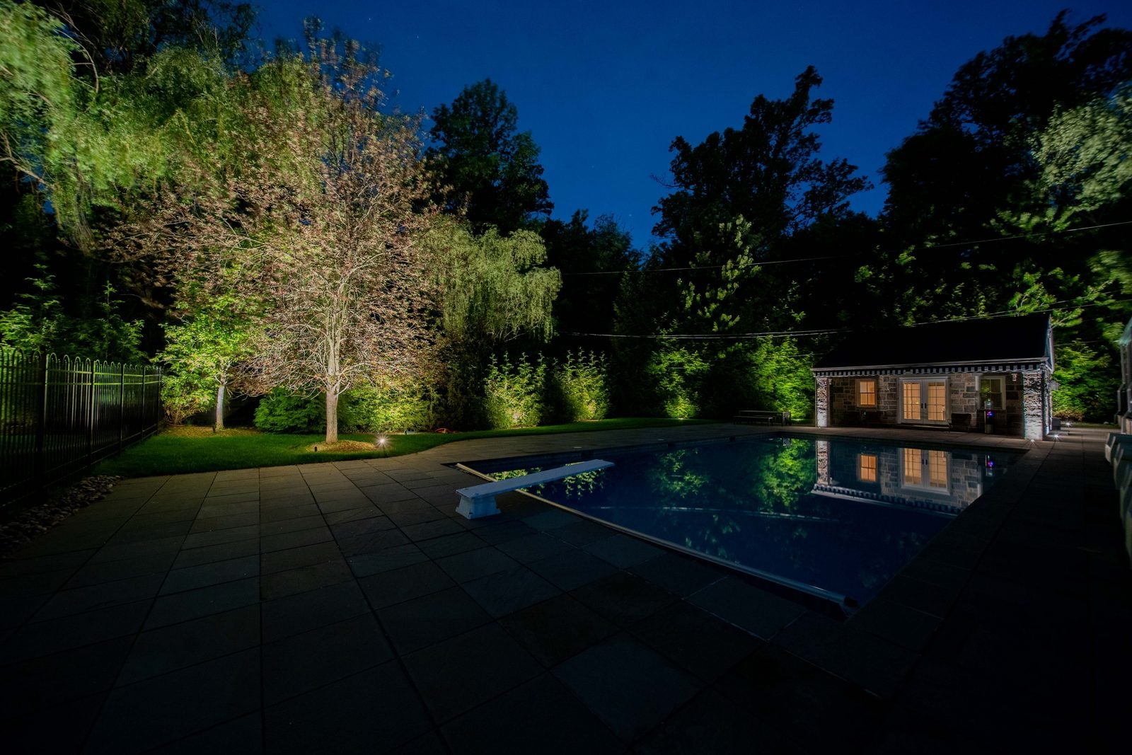 A pool and terrace lit after dark, the far planting uplit above the waterline