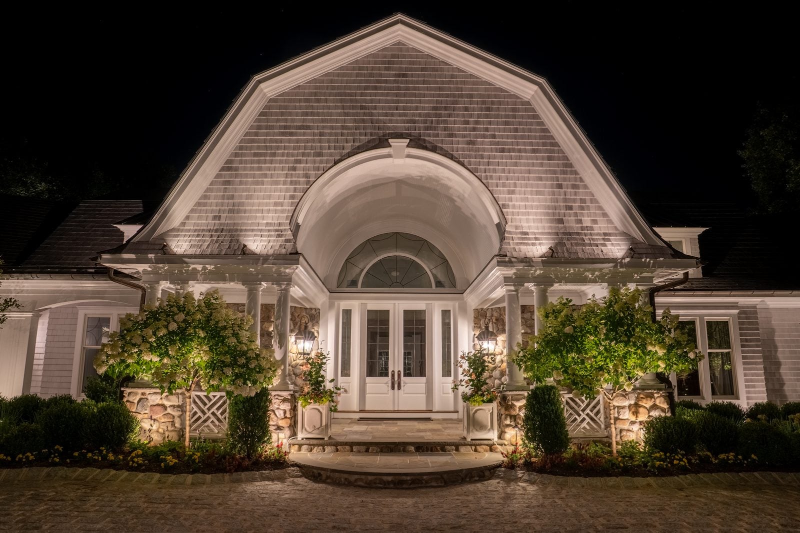 A shingle-style house at night, its tall arched window and gable lit from the ground