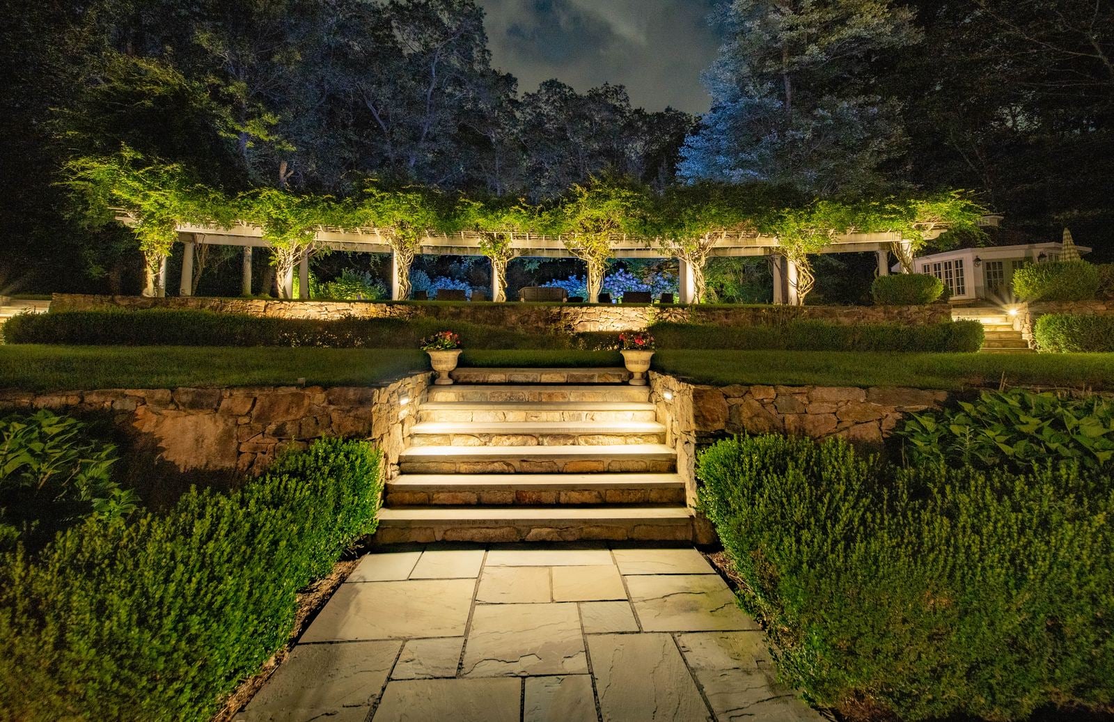 Wide stone steps climbing to a lit pergola, hedges lit low on either side
