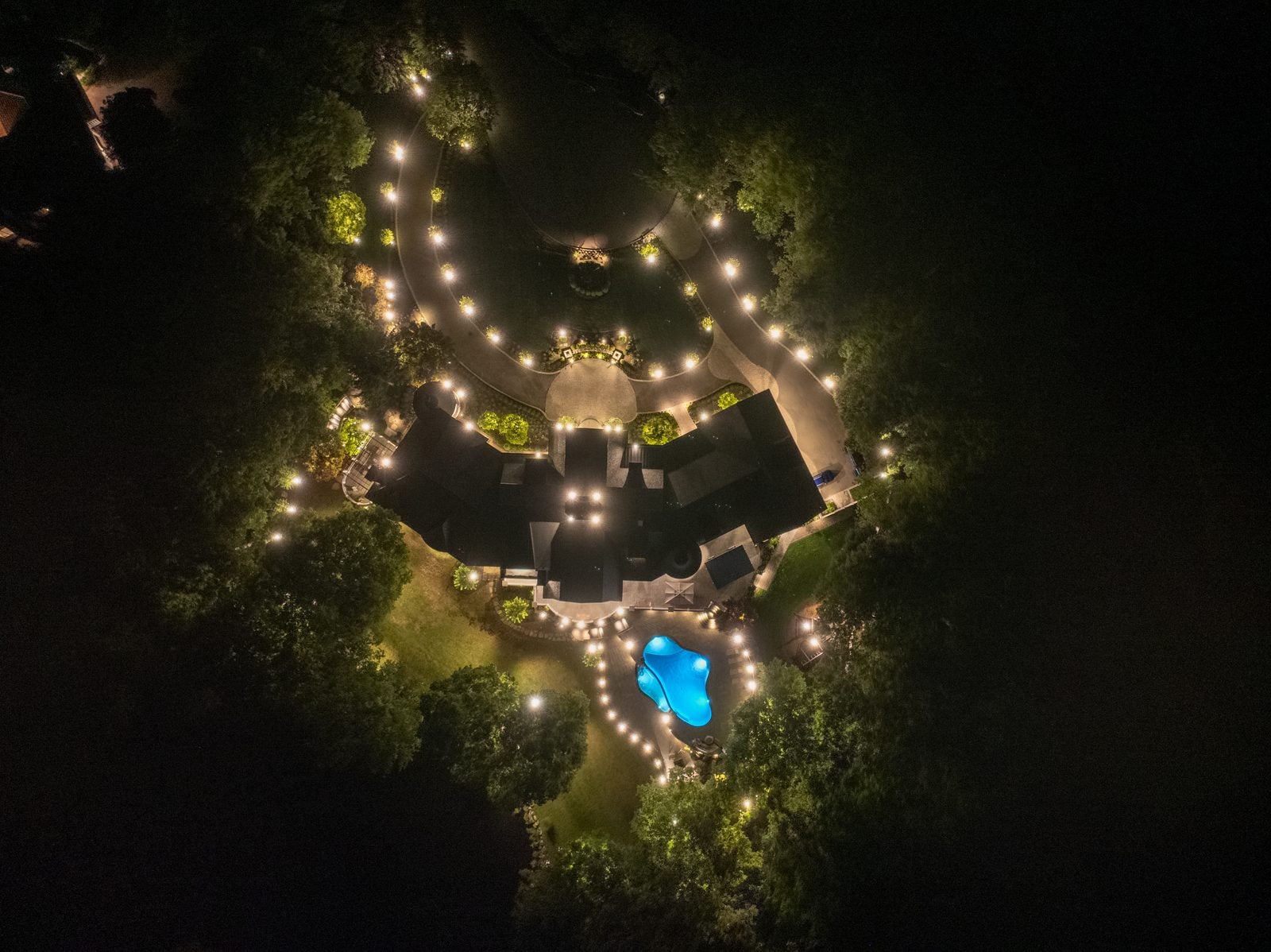 An aerial view of a property at night, warm light tracing the drive and garden beds