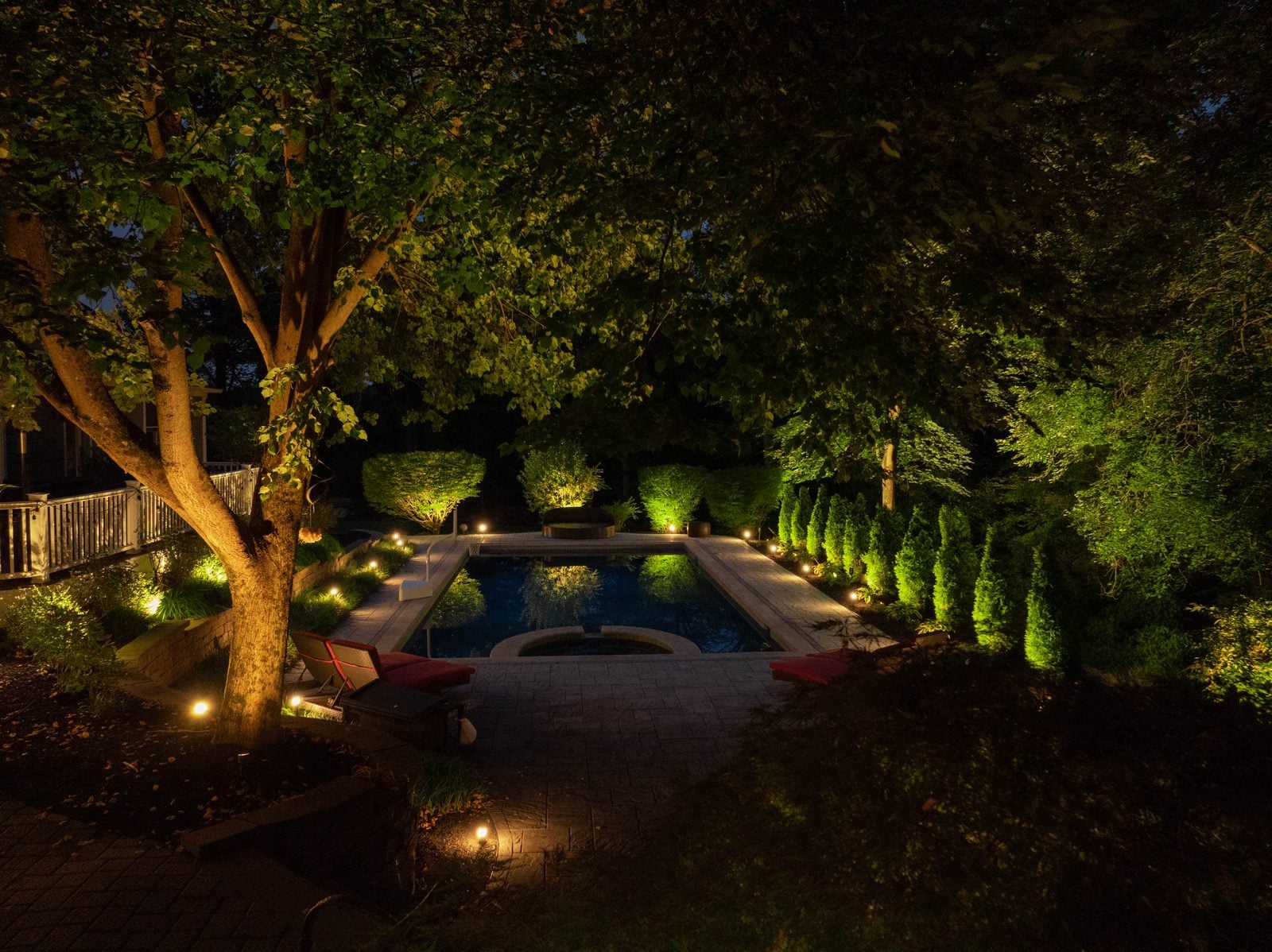A pool and terrace lit after dark, the far planting uplit above the waterline