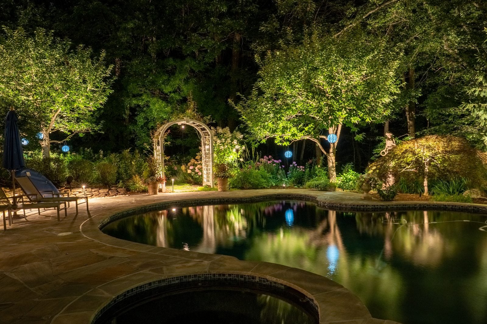 A pool and terrace lit after dark, the far planting uplit above the waterline
