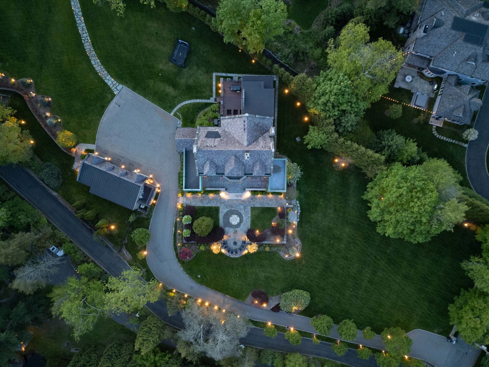 An aerial view of a property at night, warm light tracing the drive and garden beds